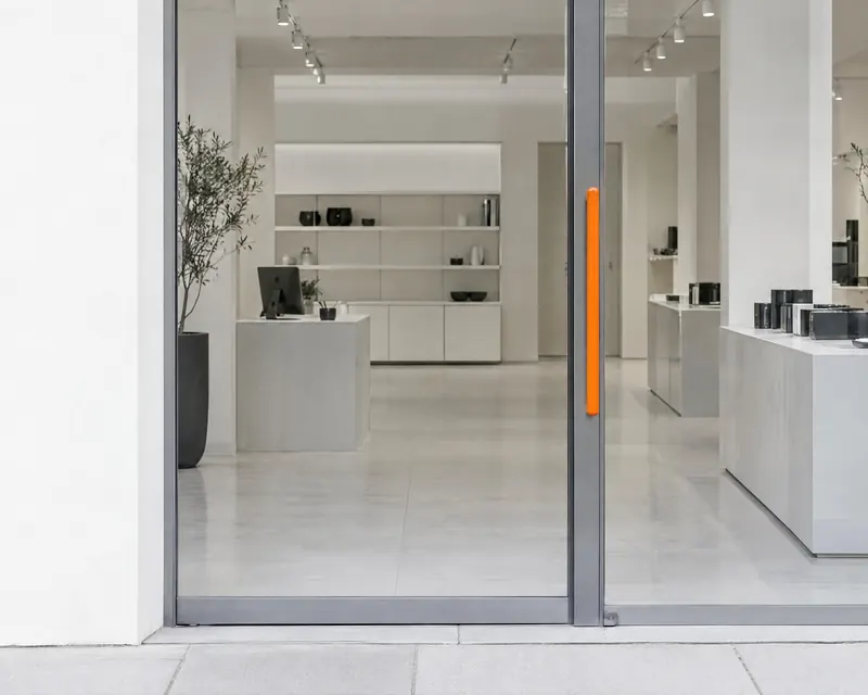 A glass shop entrance in soft daylight with an orange door pull.